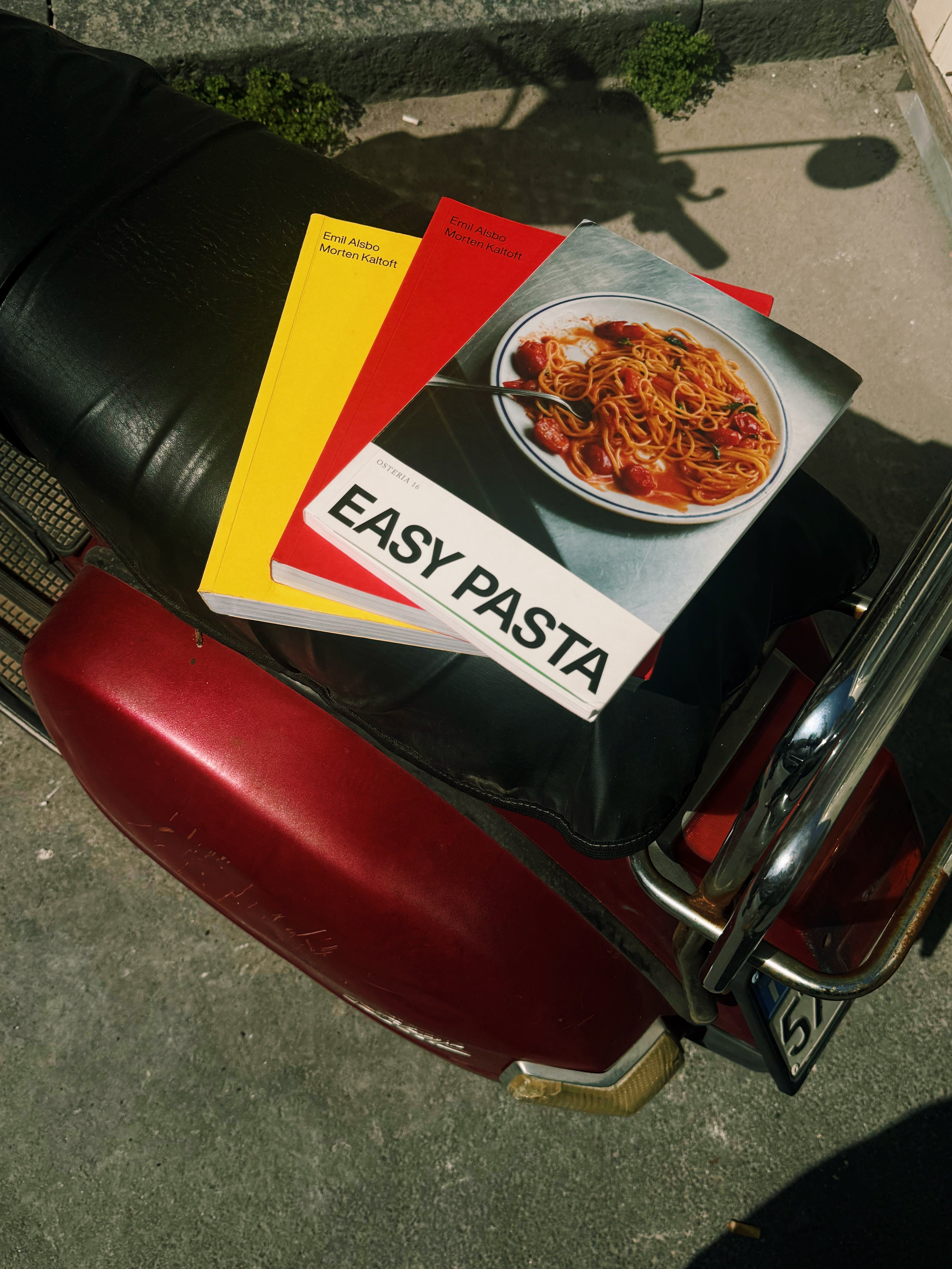 In-use: O16 kogebøger — gul og rød Osteria 16 samt Easy Pasta — på sædet af en rød Vespa-scooter, analog film udendørs | O16 cookbooks — yellow and red Osteria 16 plus Easy Pasta — on a red Vespa scooter seat, analogue film outdoors