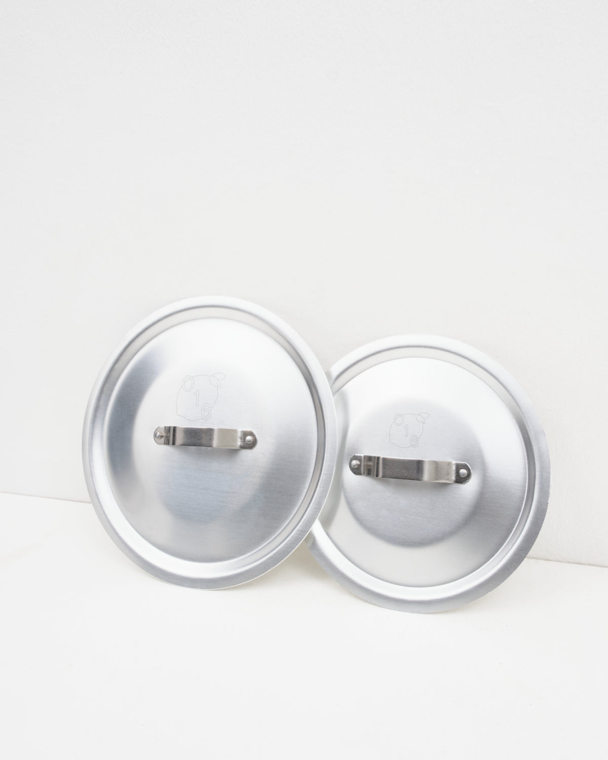 O16 to aluminiumslåg med stålgreb stillet side om side fra Osteria 16 — studieproduktbillede på hvid baggrund | O16 two aluminium lids with steel handles set side by side by Osteria 16 — studio product shot on white background