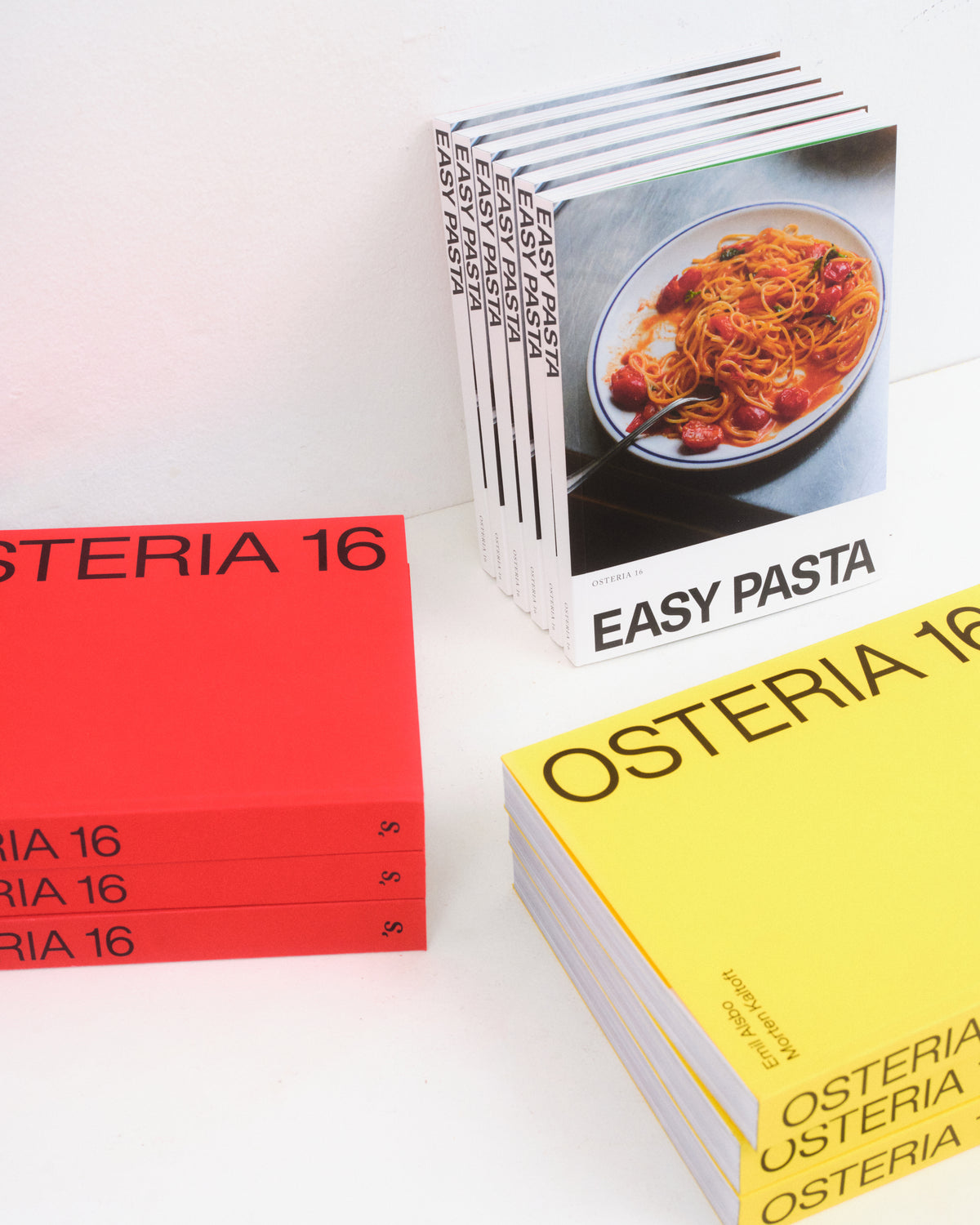O16 Osteria 16 kogebøger arrangeret — rød stabel, stående Easy Pasta og gul stabel — studie på hvid baggrund | O16 Osteria 16 cookbooks arranged — red stack, standing Easy Pasta and yellow stack — studio on white background