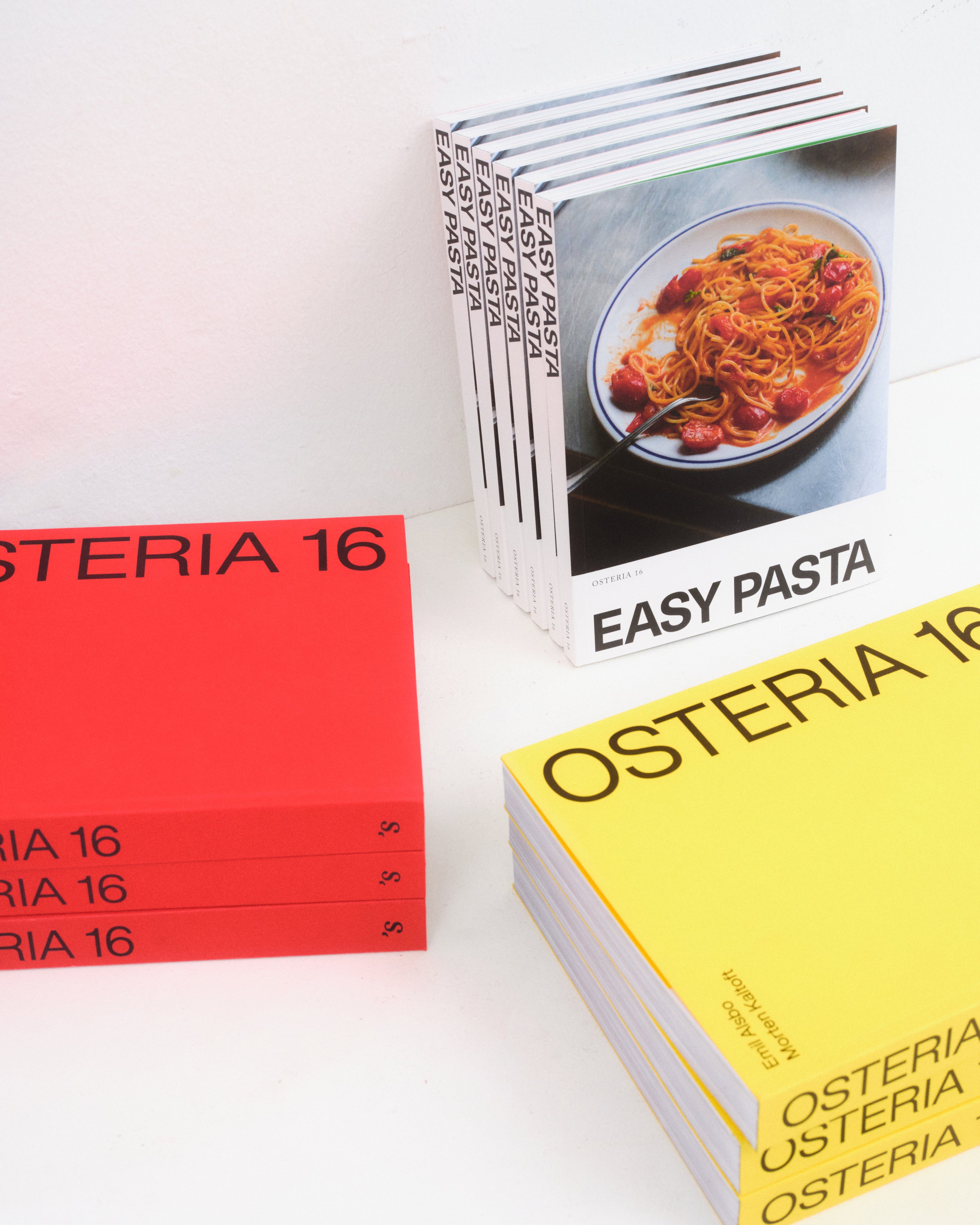 Studio: O16 Osteria 16 kogebøger arrangeret — rød stabel, stående Easy Pasta og gul stabel — studie på hvid baggrund | O16 Osteria 16 cookbooks arranged — red stack, standing Easy Pasta and yellow stack — studio on white background
