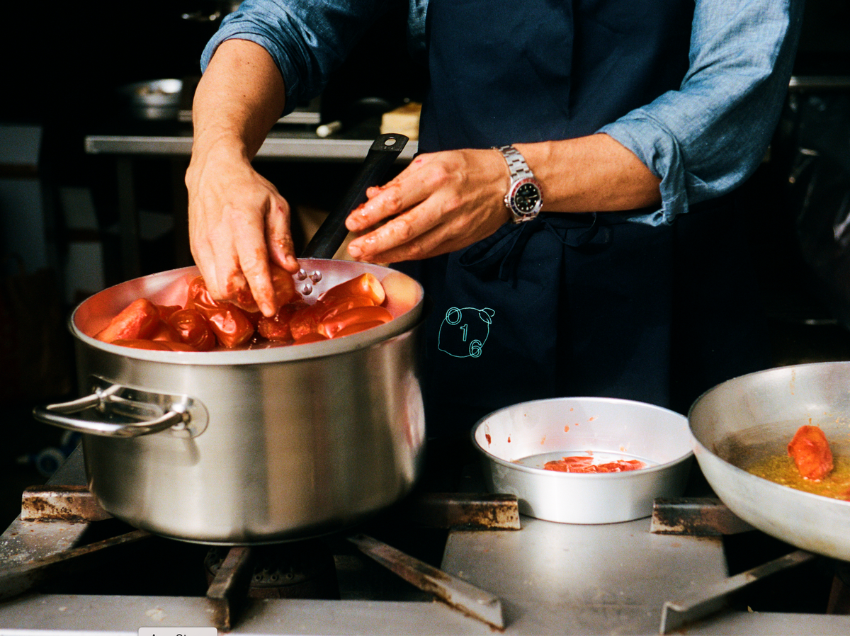 Kok presser friske tomater i en stor gryde på komfuret, iført navy O16-forklæde, analog film, Osteria 16-køkken | Chef pressing fresh tomatoes in a large stockpot on the stove, wearing a navy O16 apron, analogue film, Osteria 16 kitchen