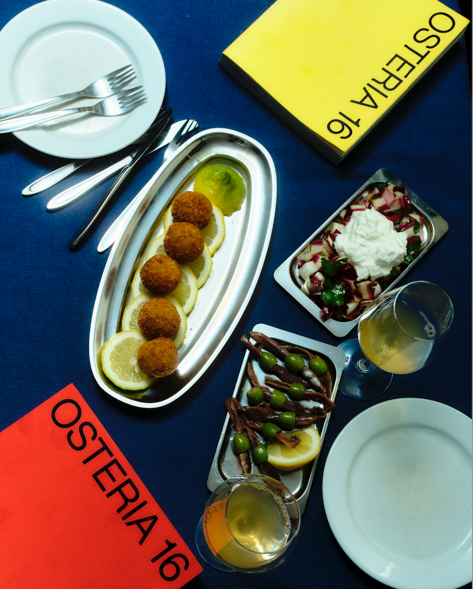 Osteria 16 kogebøger (gul og rød udgave) med arancini på citron, ansjoser med oliven, burrata-salat og hvidvin, redaktionel flat-lay | Osteria 16 cookbooks (yellow and red editions) with arancini on lemon, anchovies and olives, burrata salad and white wine, editorial flat-lay