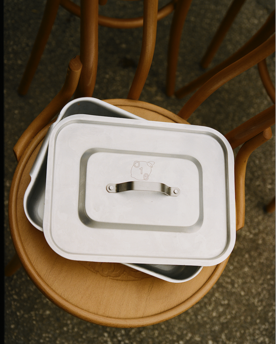 O16-aluminiumsfad med låg og indgraveret O16-grisemærke stablet på en wienerstol udendørs | O16 aluminium lidded box engraved with the O16 pig mark, stacked on a bentwood chair outdoors — Osteria 16