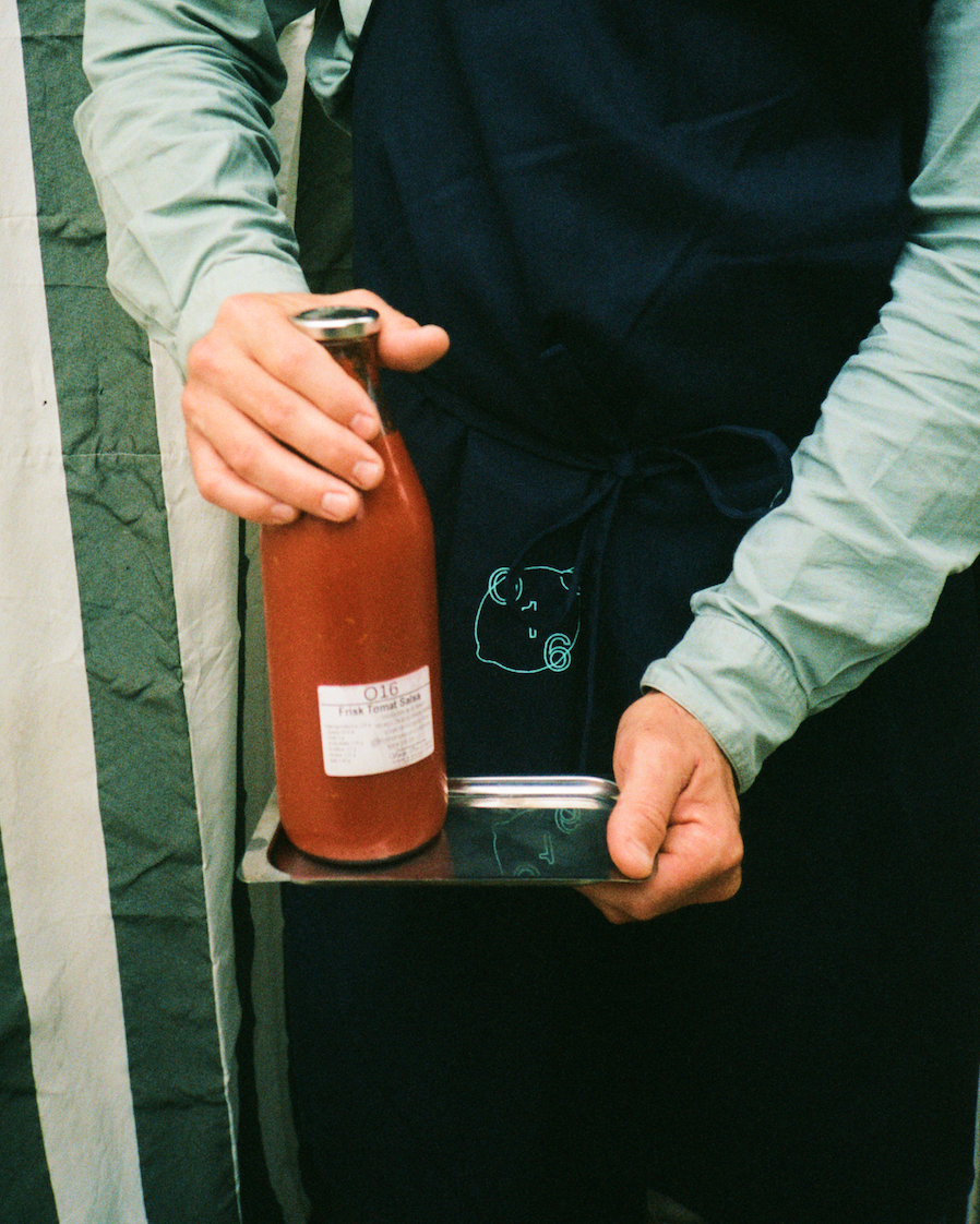Person i forklæde med O16 grislogo holder flaske O16 tomatsauce på stålfad — Osteria 16 passata | Person in an apron with the O16 pig logo holding a bottle of O16 tomato sauce on a steel tray — Osteria 16 passata