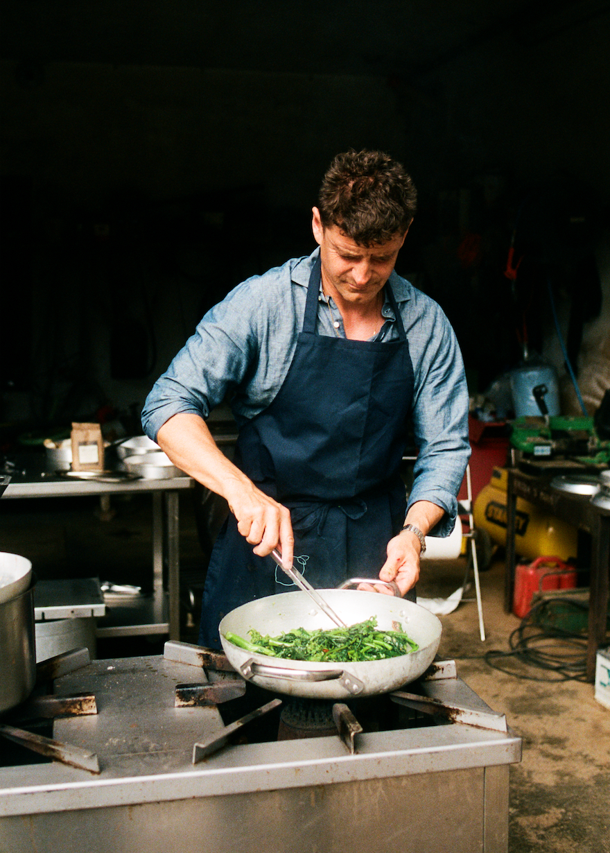 In-use Osteria 16 kok i blåt forklæde tilbereder grønne blade i stor stålpande over gasblus, rustikt køkken, analog film | Osteria 16 chef in a blue apron cooking leafy greens in a large steel pan over a gas flame, rustic kitchen, analogue film