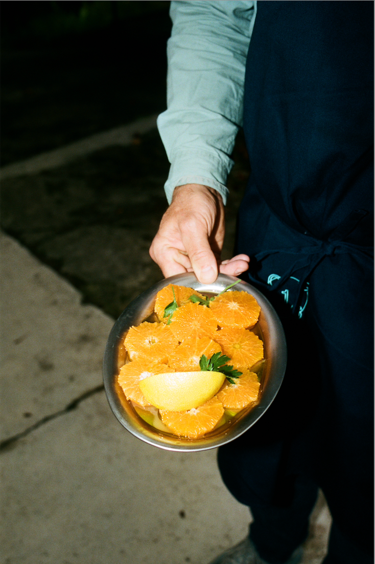 In-use: Osteria 16 kok holder et stålfad med appelsinskiver, persille og citron udendørs i aftenlys, analog film | Osteria 16 chef holding a steel dish of sliced oranges, parsley and lemon outdoors in evening light, analogue film