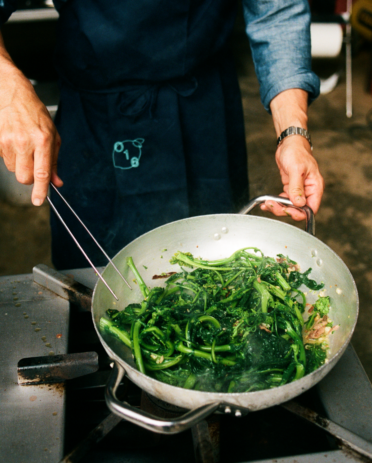 Kok vender broccolini i en aluminiumspande med O16 Pinze køkkentang, iført navy O16-forklæde, analog film | Chef tossing broccoli rabe in an aluminium pan with O16 Pinze kitchen tongs, wearing a navy O16 apron, analogue film
