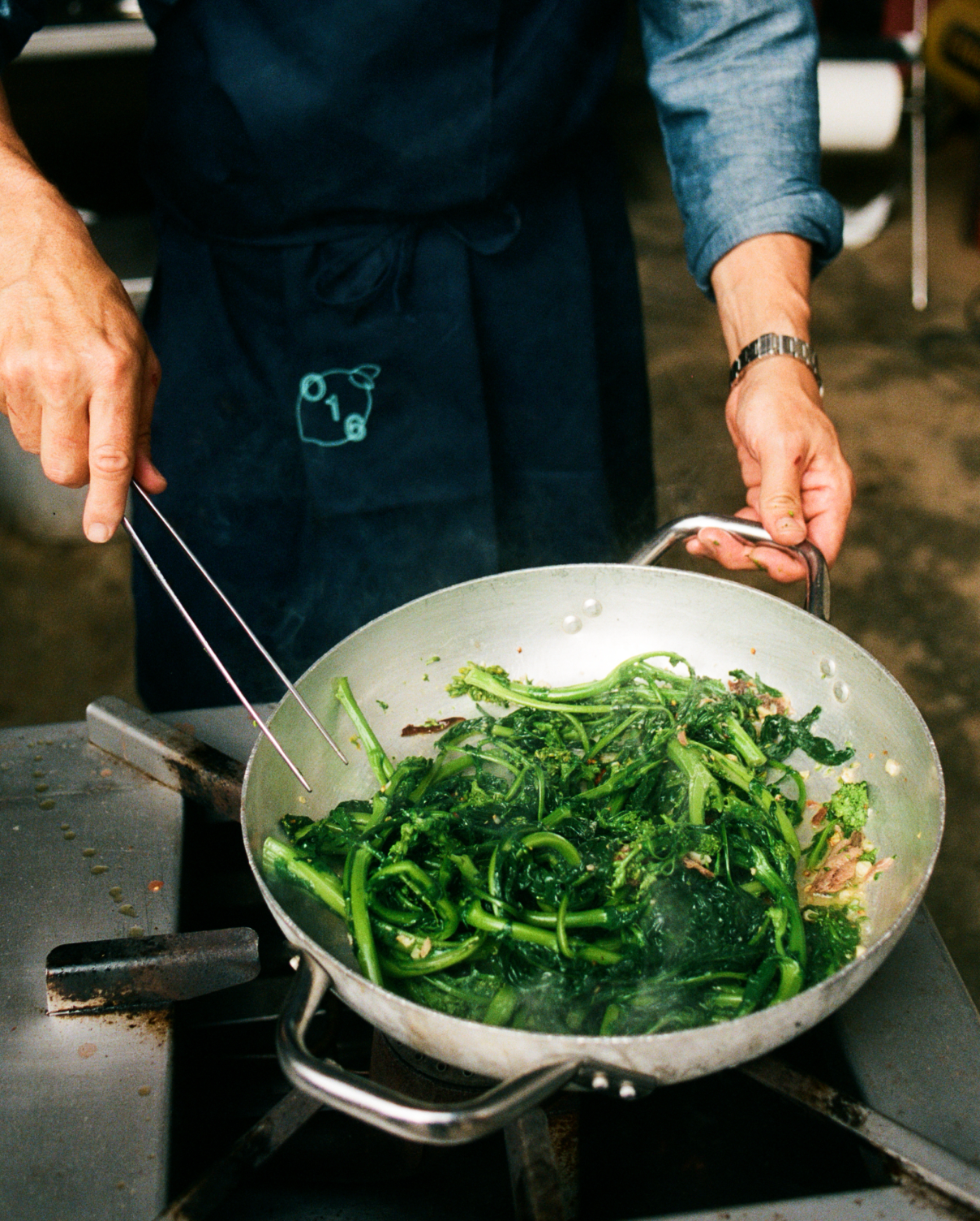In-use: Kok vender broccolini i en aluminiumspande med O16 Pinze køkkentang, iført navy O16-forklæde, analog film | Chef tossing broccoli rabe in an aluminium pan with O16 Pinze kitchen tongs, wearing a navy O16 apron, analogue film