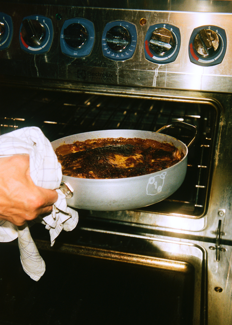En hånd med viskestykke tager en O16-aluminiumsbradepande med en bagt ret ud af en Electrolux-ovn | A cloth-wrapped hand pulling an O16 aluminium roasting pan with a baked dish from an Electrolux oven — Osteria 16