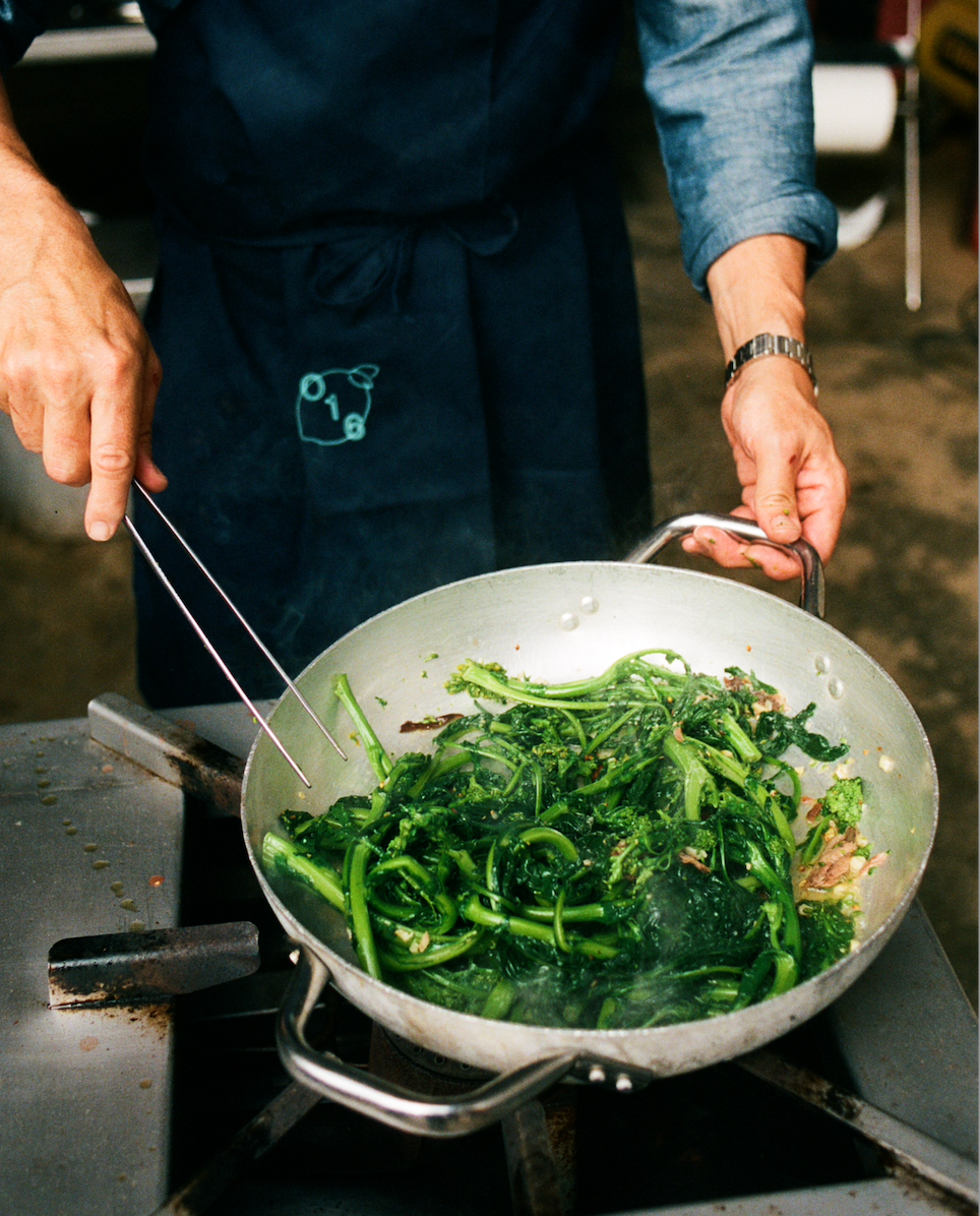 In-use: Kok i O16-forklæde sauterer cime di rapa (broccoligrønt) i en O16-aluminiumspande på gasblus | Cook in an O16 apron sautéing cime di rapa greens in an O16 aluminium pan on a gas ring — Osteria 16