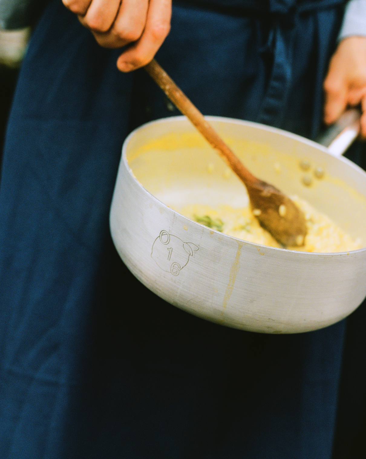 In-use: Kok i mørkeblåt forklæde rører risotto med en træske i en O16-aluminiumsgryde med indgraveret O16-grisemærke | Cook in a navy apron stirring risotto with a wooden spoon in an O16 aluminium saucepan engraved with the O16 pig mark — Osteria 16