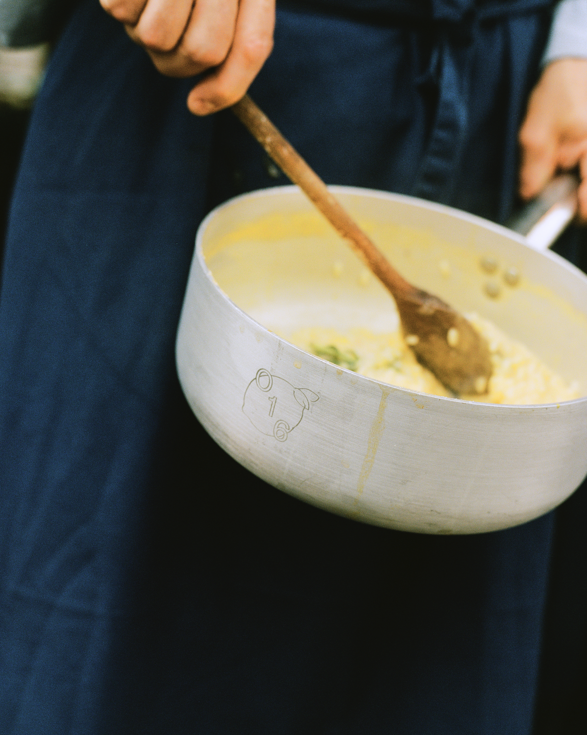 In-use: Kok i mørkeblåt forklæde rører risotto med en træske i en O16-aluminiumsgryde med indgraveret O16-grisemærke | Cook in a navy apron stirring risotto with a wooden spoon in an O16 aluminium saucepan engraved with the O16 pig mark — Osteria 16