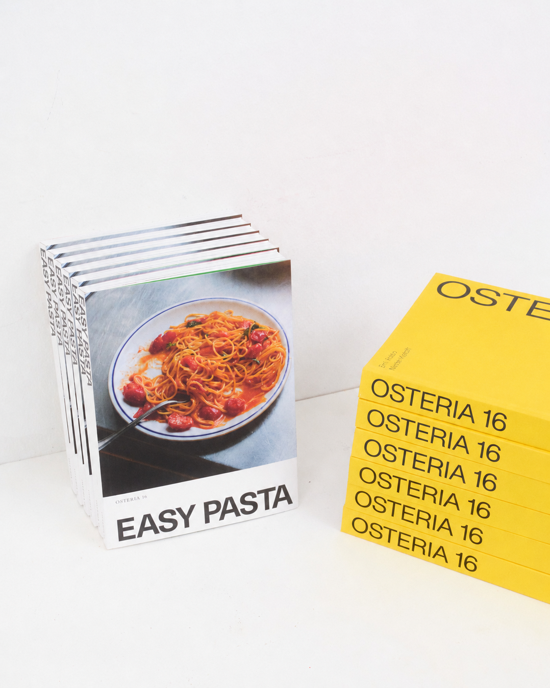 Studio: O16 Osteria 16 kogebøger — stående stabel Easy Pasta ved siden af liggende gul Osteria 16 — studie på hvid baggrund | O16 Osteria 16 cookbooks — standing Easy Pasta stack beside a lying yellow Osteria 16 stack — studio on white background