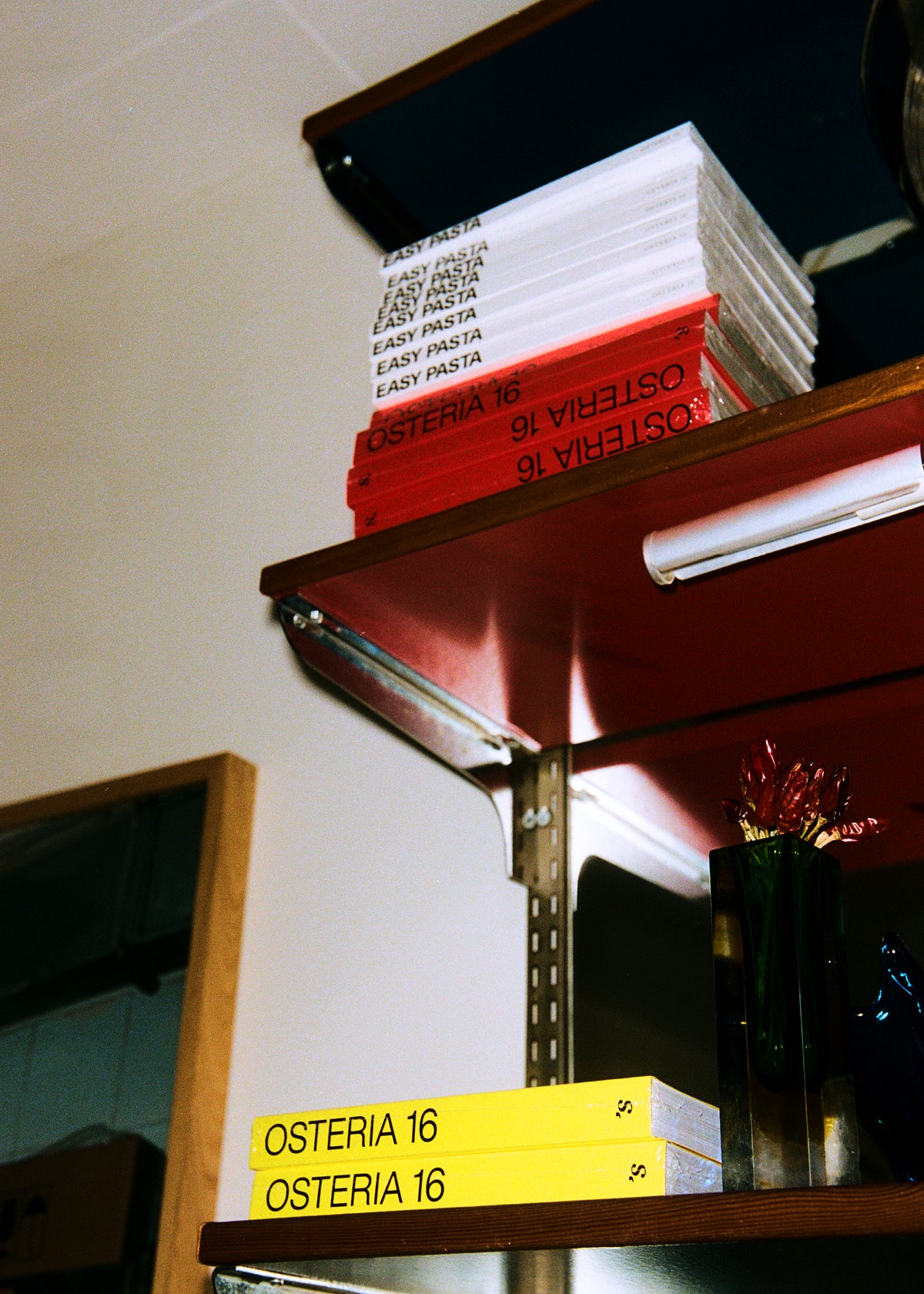 O16 kogebøger på hylde — Easy Pasta og røde Osteria 16 øverst, gule Osteria 16 nedenfor — analog film i butiksmiljø | O16 cookbooks on a shelf — Easy Pasta and red Osteria 16 above, yellow Osteria 16 below — analogue film in-store