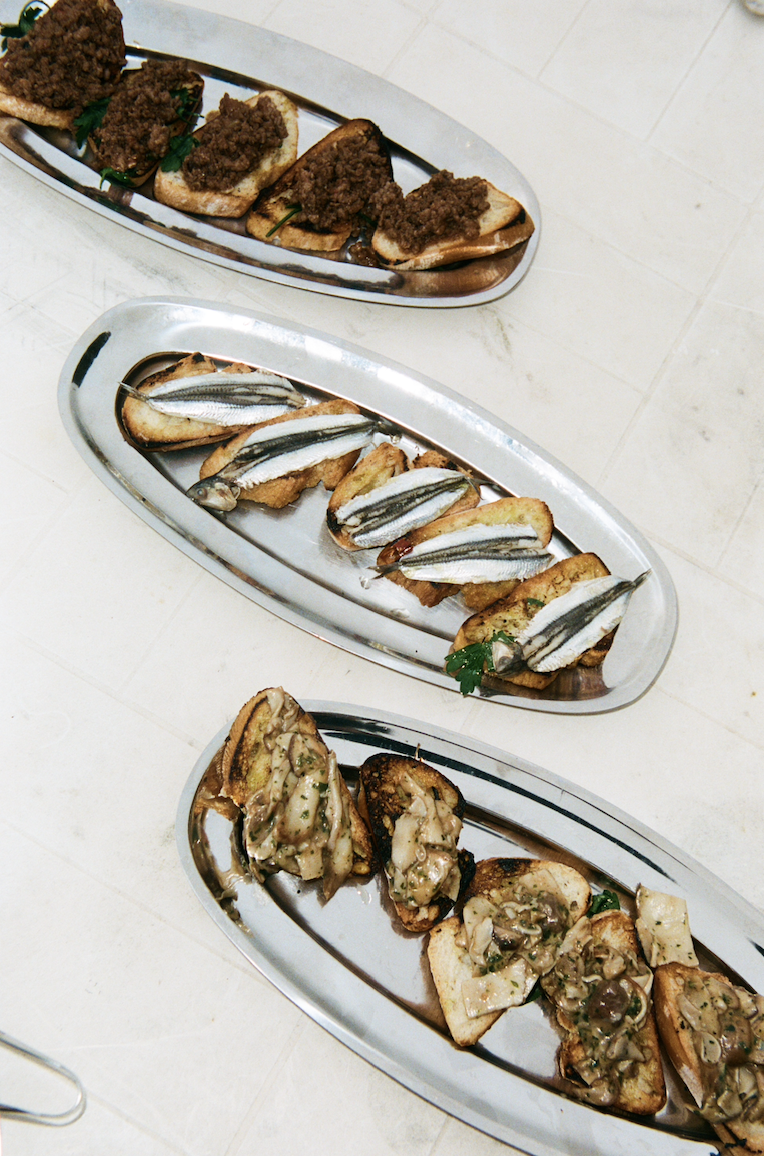 In-use: Osteria 16 crostini på tre ovale stål-serveringsfade — ansjos, oliventapenade og svampe — på marmorgulv, analog film | Osteria 16 crostini on three oval steel serving trays — anchovy, olive tapenade and mushroom — on a marble floor, analogue film