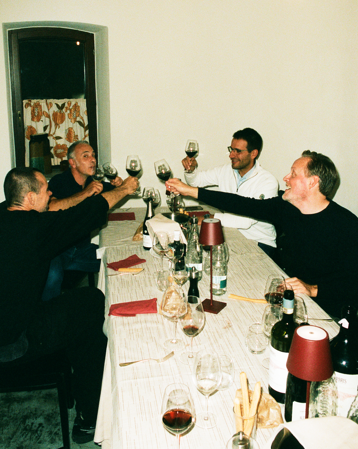 In-use: Analogt aftenfoto: fire mænd skåler i rødvin ved langbord i italiensk osteria; vinflasker og bordlamper. | Analogue evening photo: four men toasting red wine at a long table in an Italian osteria; wine bottles and table lamps.