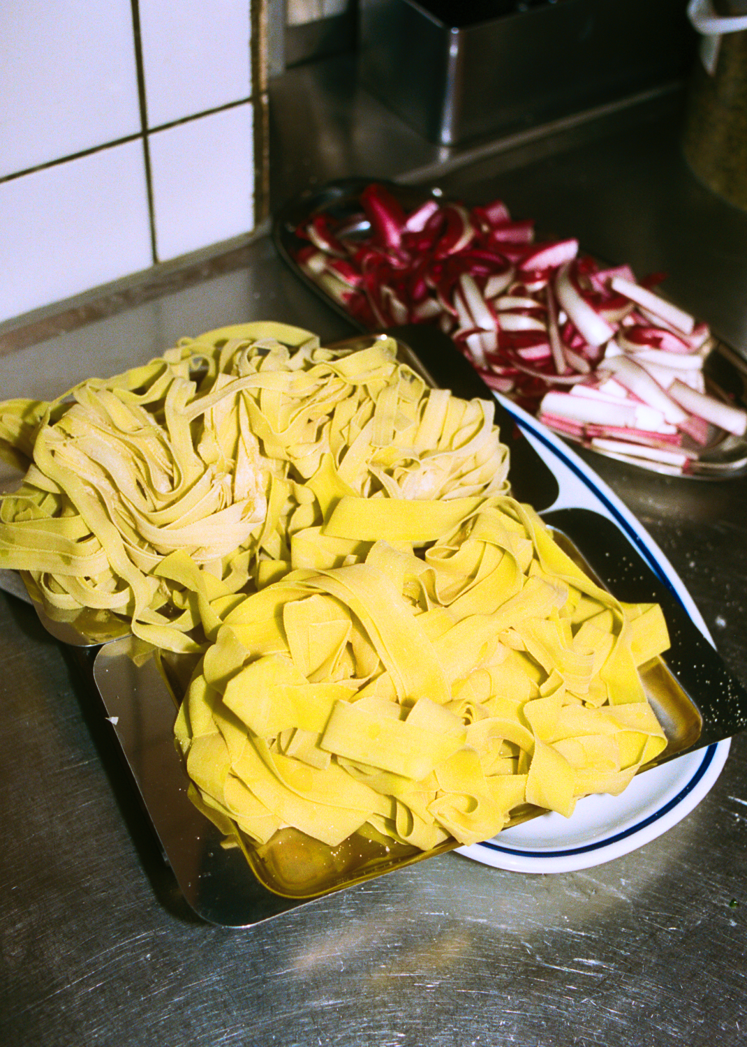 In-use: Frisk hjemmelavet tagliatelle på O16 stålfad ved køkkenbord, snittet radicchio bagved — Osteria 16 køkkenfoto | Fresh house-made tagliatelle on an O16 steel tray by the kitchen counter, sliced radicchio behind — Osteria 16 kitchen shot