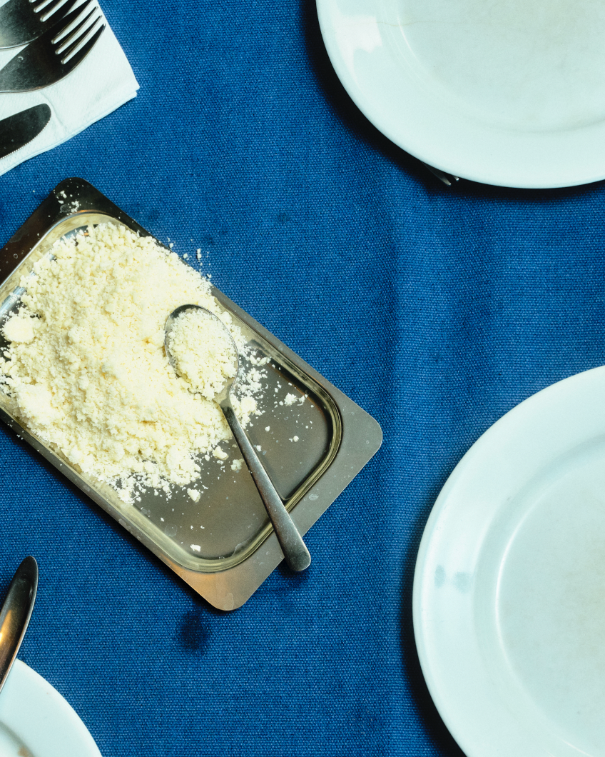 In-use: Revet parmesan i O16 stålfad med ske på blå dug, hvide tallerkener rundt om — rustikt Osteria 16 bordfoto | Grated Parmesan in an O16 steel tray with a spoon on blue linen, white plates around — rustic Osteria 16 table shot