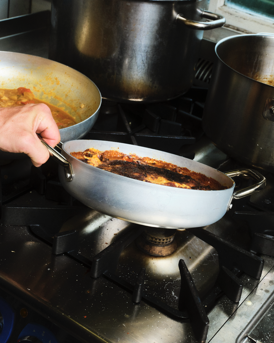 In-use Hånd holder et ovalt aluminiumsfad med braiseret ret over gasblus i Osteria 16-køkkenet, analog film | Hand holding an oval aluminium dish with a braised dish over a gas flame in the Osteria 16 kitchen, analogue film