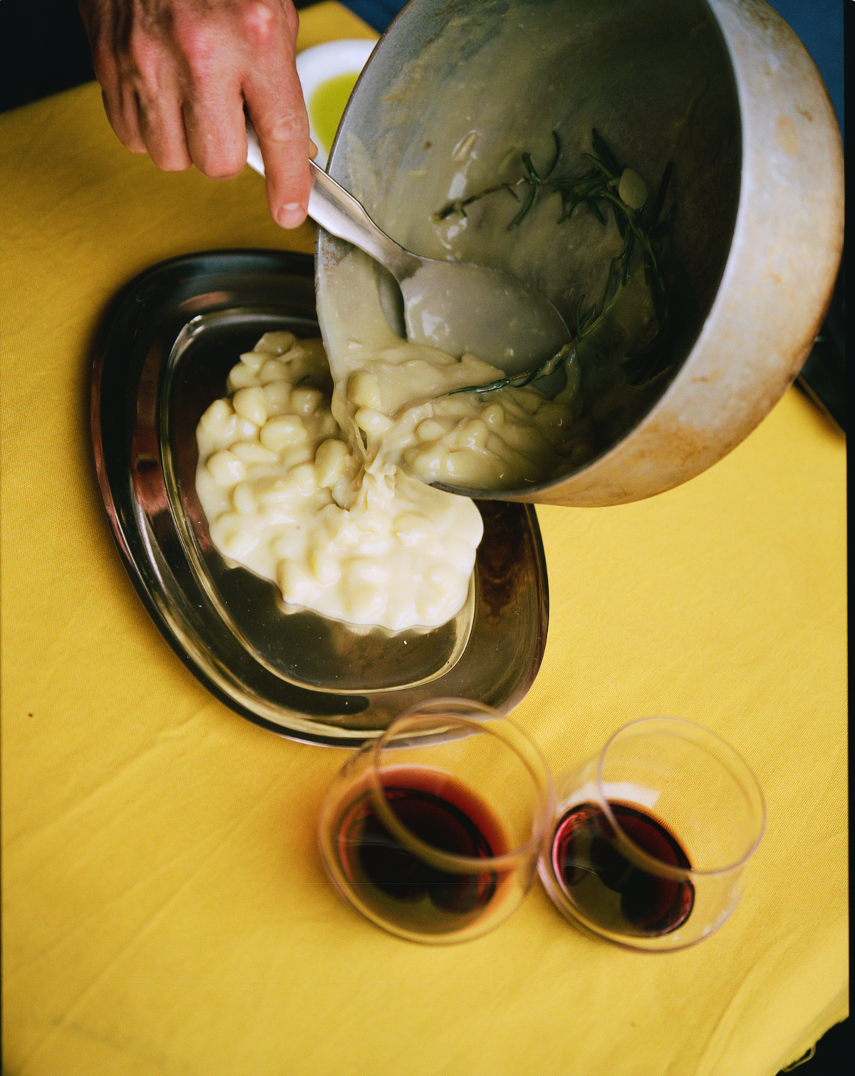 Hånd øser gnocchi i flødesauce af O16 aluminiumsgryde over ovalt stål-vassoio; gul dug og rødvin. | Hand serving gnocchi in cream sauce from an O16 aluminium pot onto an oval stainless vassoio; yellow cloth, red wine.