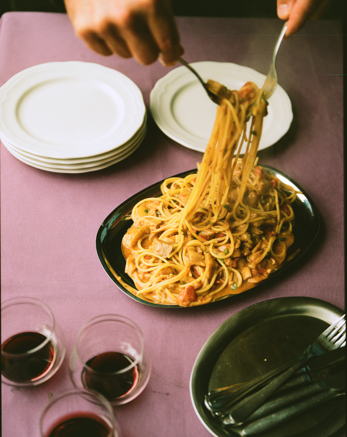 Hænder løfter spaghetti fra mørkt ovalt O16-vassoio; lilla dug, stablede tallerkener og rødvin, Osteria 16. | Hands lifting spaghetti from a dark oval O16 vassoio; purple cloth, stacked plates and red wine, Osteria 16.