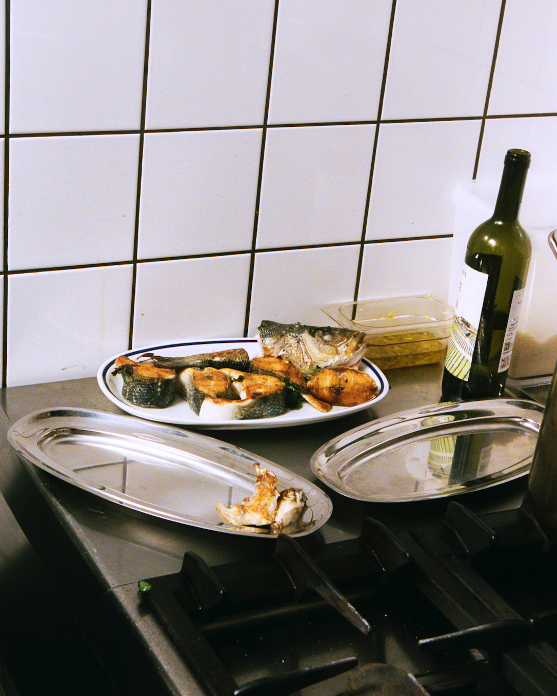 In-use: Osteria 16-køkken — grillet fisk på et hvidt ovalt fad, stål-serveringsfade, vinflaske og hvide fliser, analog film | Osteria 16 kitchen — grilled fish on a white oval plate, steel serving trays, a wine bottle and white tiles, analogue film