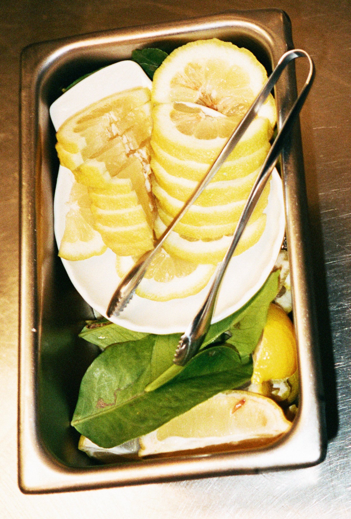 O16 Pinze køkkentang i stål i gastrobakke med citronskiver og laurbær — italiensk køkken i brug | O16 Pinze stainless steel kitchen tongs in a gastro tray with lemon slices and bay leaves — Italian kitchen in use