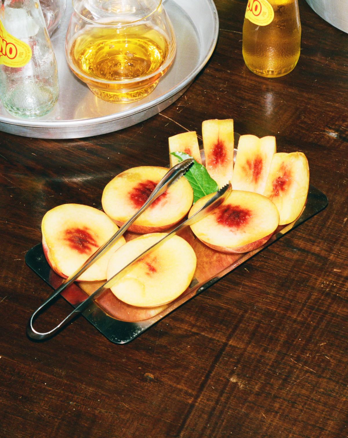 O16 Pinze køkkentang i stål med friske ferskenskiver på fad — lifestyle shot ved bordet | O16 Pinze stainless steel kitchen tongs with fresh sliced peaches on a tray — lifestyle table shot