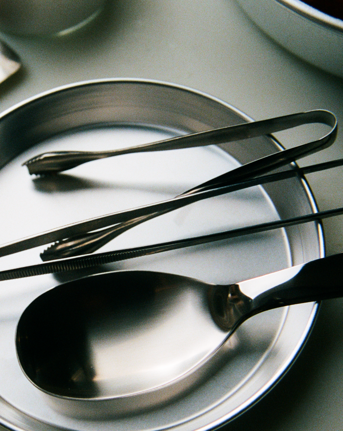 O16 Pinze serveringstang i stål med serveringsske på stålfade — nærbillede af form og finish | O16 Pinze stainless steel serving tongs with a serving spoon on steel plates — close-up of form and finish