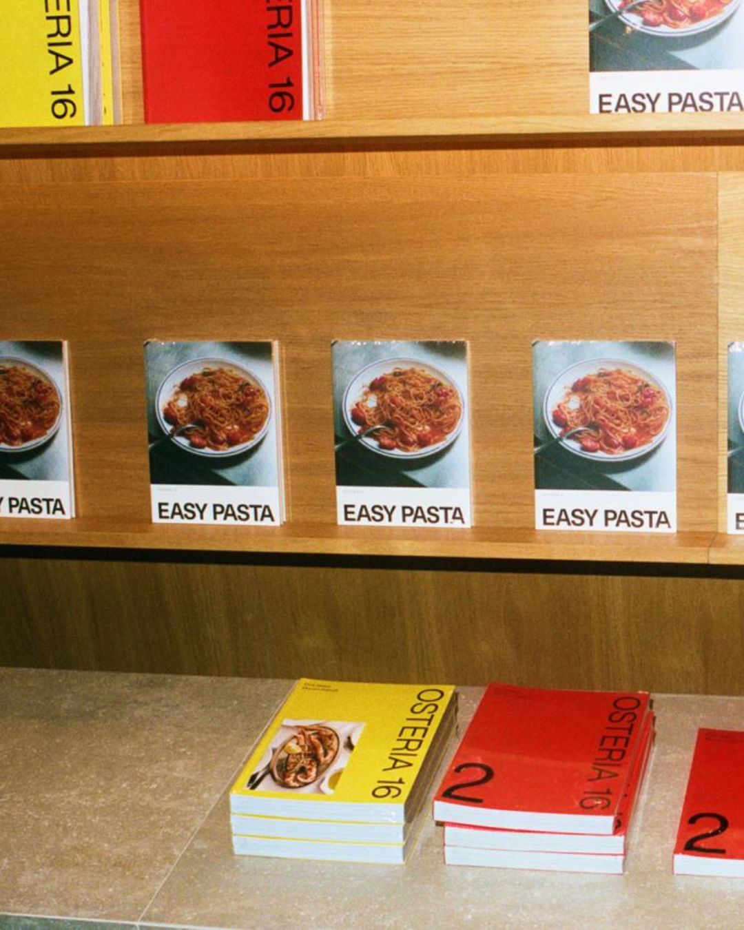 O16 butiksdisplay med Easy Pasta-forsider på træhylde og gule og røde Osteria 16 kogebøger på disken, analog film | O16 retail display with Easy Pasta covers on a wooden shelf and yellow and red Osteria 16 cookbooks on the counter, analogue film