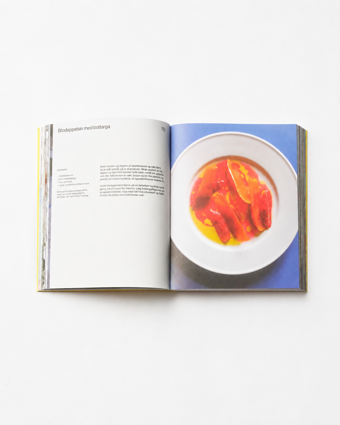 !Pages: Opslag i O16 kogebog med Blodappelsin med bottarga-opskriften og foto af blodappelsin i olie på blåt bord. | Spread in the O16 cookbook showing the Blood orange with bottarga recipe, blood orange in oil on a blue table.