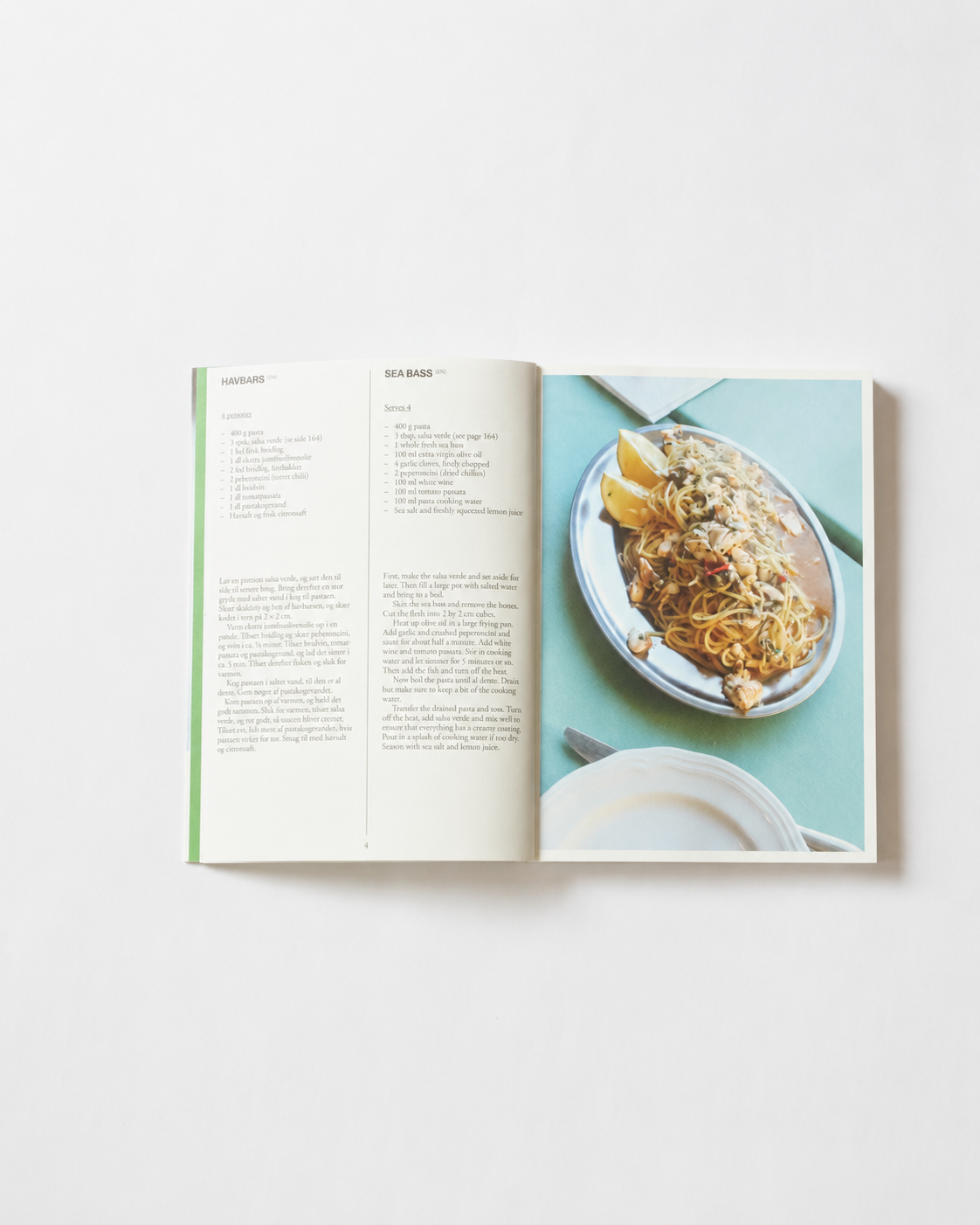 !Pages: Opslag i O16 kogebog med Havbars, Sea Bass, og foto af spaghetti med havbars på turkis bord. | Spread in the O16 cookbook showing the Sea Bass recipe with a photo of spaghetti and sea bass on a turquoise table.
