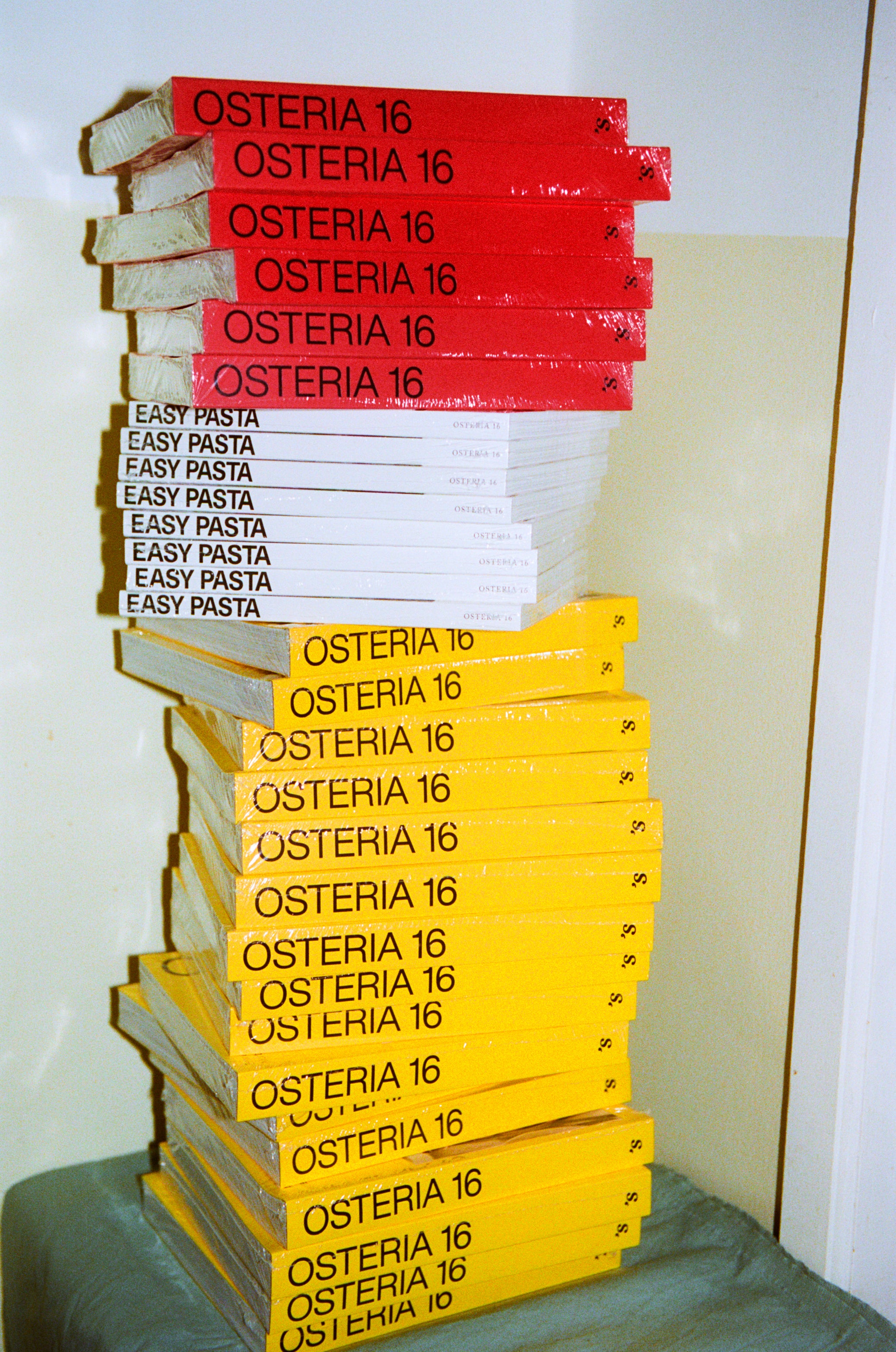 In-use: Stabel af O16 kogebøger — røde og gule Osteria 16 samt hvide Easy Pasta — analog film, rustik italiensk stemning | Stack of O16 cookbooks — red and yellow Osteria 16 plus white Easy Pasta — analogue film, rustic Italian mood