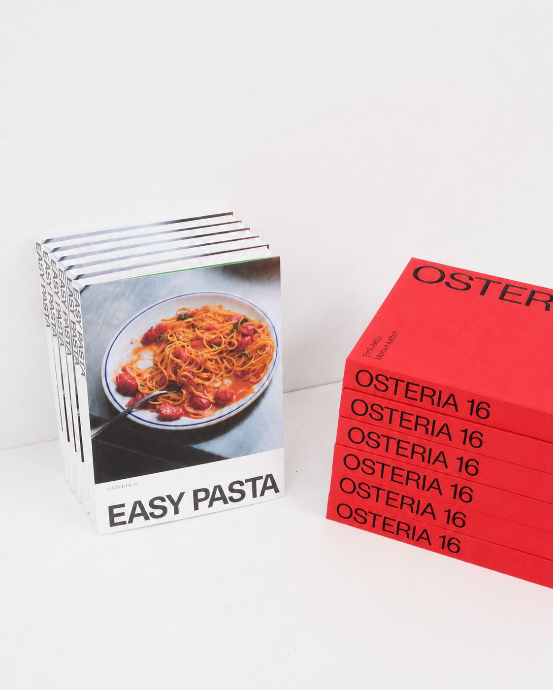 Studio: Stak af "Easy Pasta" kogebøger ved siden af stak røde Osteria 16 bøger på hvid baggrund — studieoptagelse | Stack of "Easy Pasta" cookbooks beside a stack of red Osteria 16 books on white — studio shot