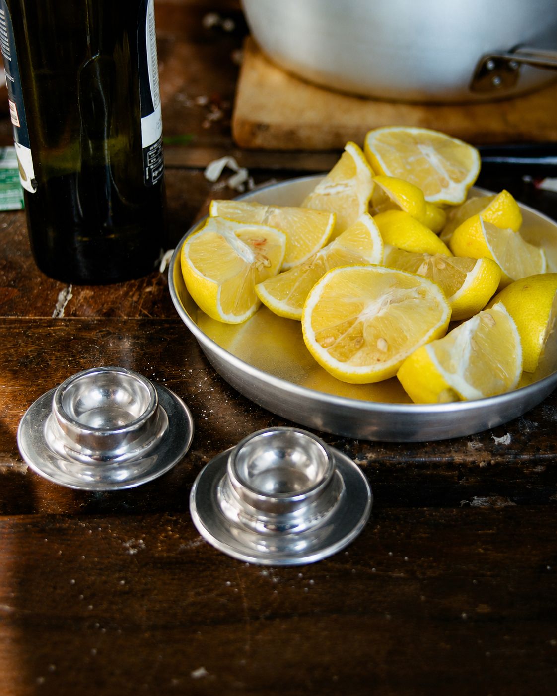 O16 stålfad med udpressede citronhalvdele på rustikt træbord, vinflaske og to tinholdere; køkkenforberedelse. | O16 stainless plate of squeezed lemon halves on a rustic wood table, wine bottle and two pewter holders; kitchen prep.