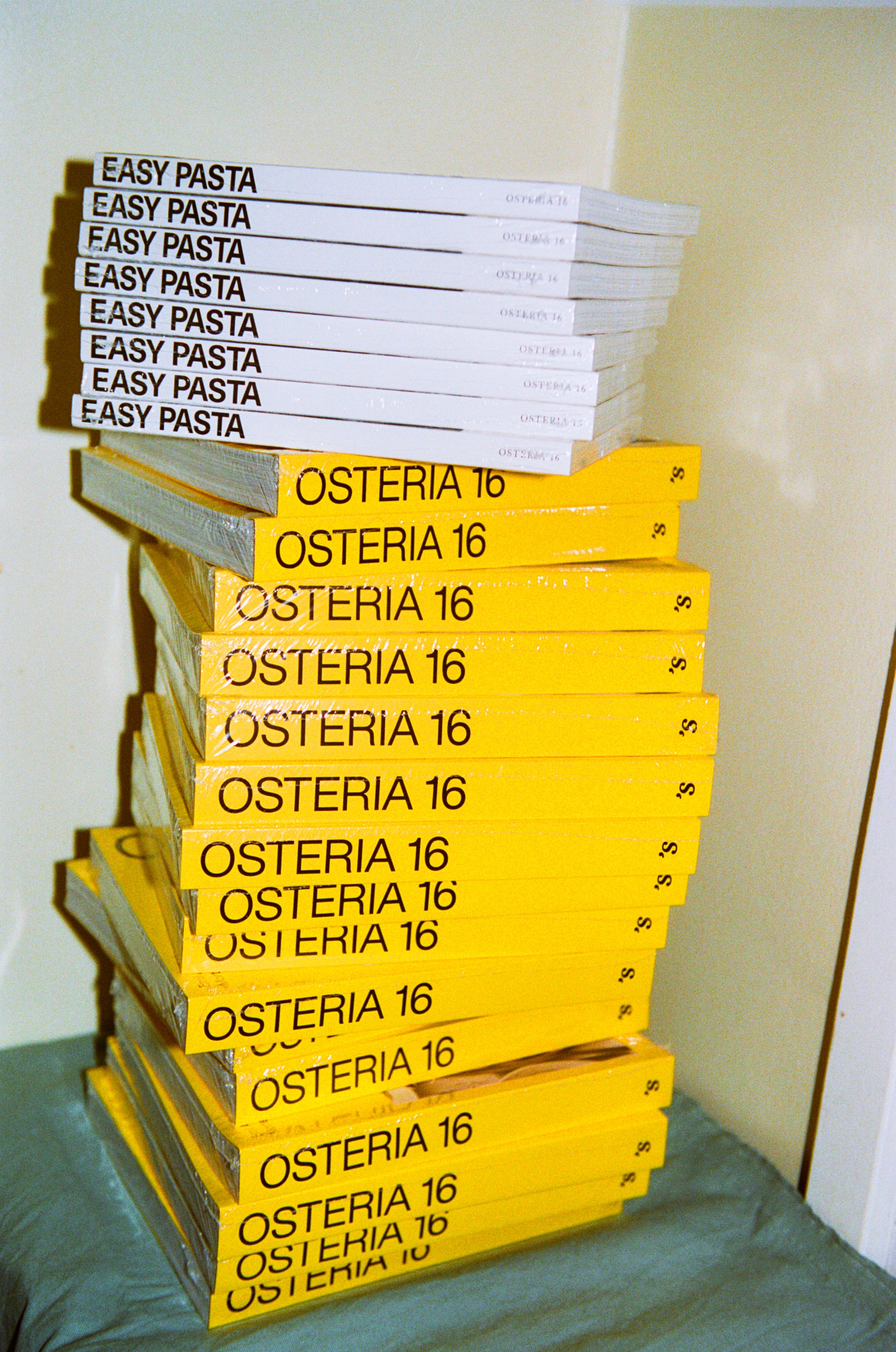 In-use: Høj stak Osteria 16 'Easy Pasta' hæfter og gule Osteria 16 kogebøger i et hjørne, film-foto | Tall stack of Osteria 16 'Easy Pasta' booklets and yellow Osteria 16 cookbooks in a corner, film photo