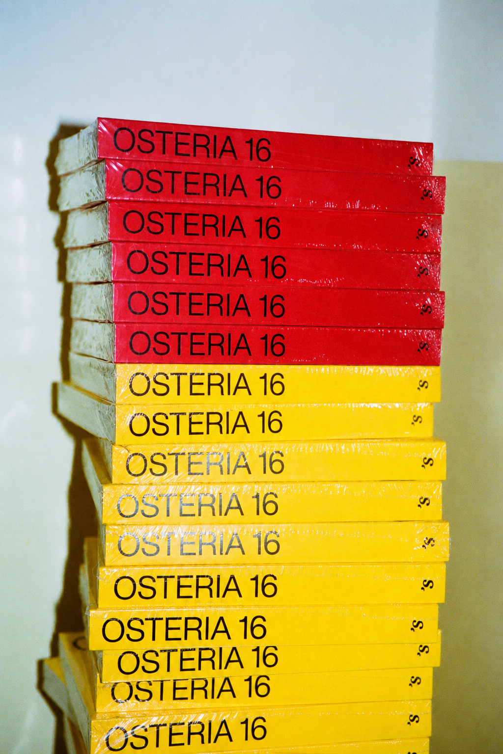 Høj stabel af indpakkede O16 Osteria 16 kogebøger, røde øverst og gule nedenfor, analog film i butik | Tall stack of shrink-wrapped O16 Osteria 16 cookbooks, red on top and yellow below, analogue film in-store
