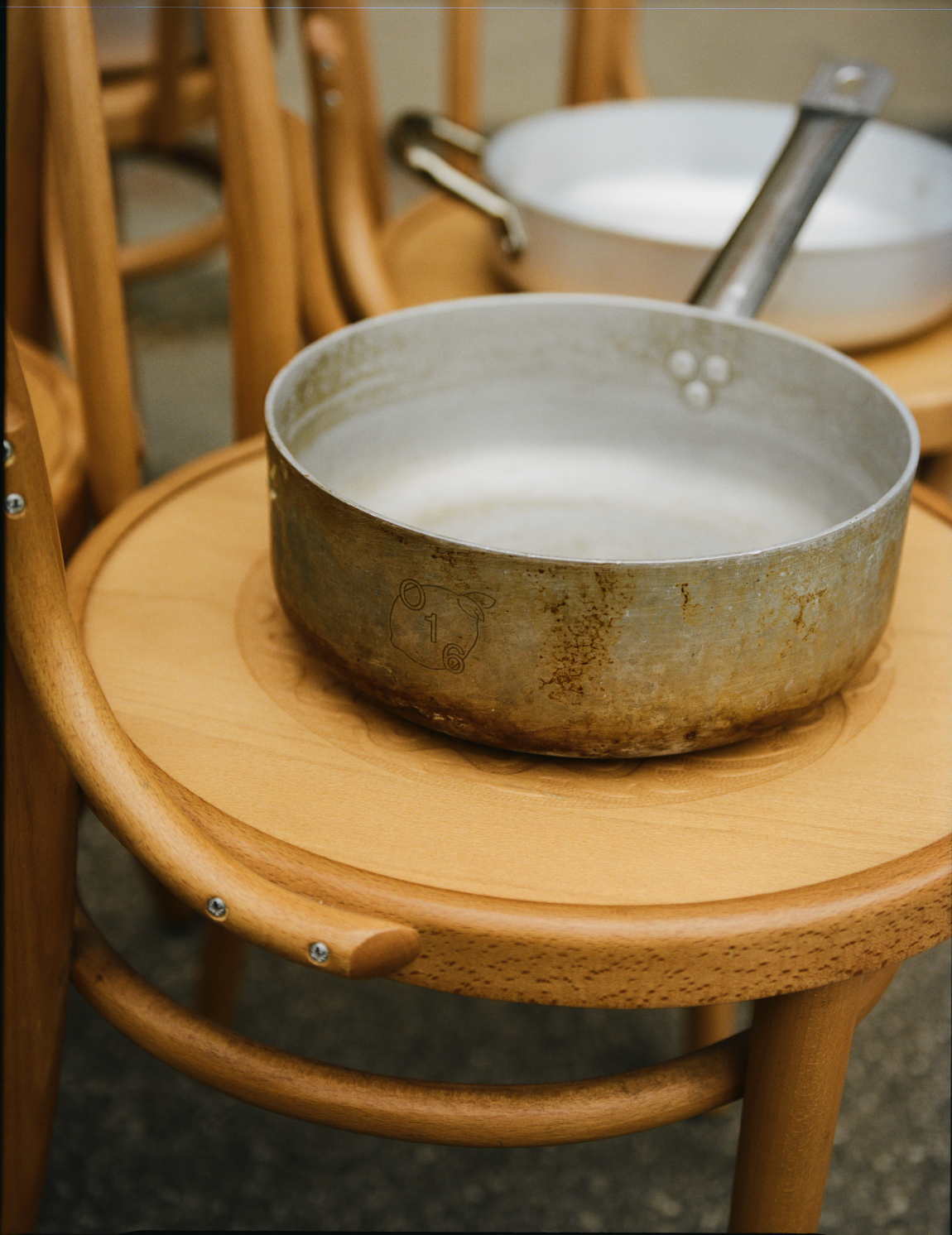 To rustikke O16 aluminiums-casseruola-gryder med mel står på bøjletræsstole i køkkenet hos Osteria 16. | Two rustic O16 aluminium casseruola pots dusted with flour rest on bentwood café chairs in the Osteria 16 kitchen.