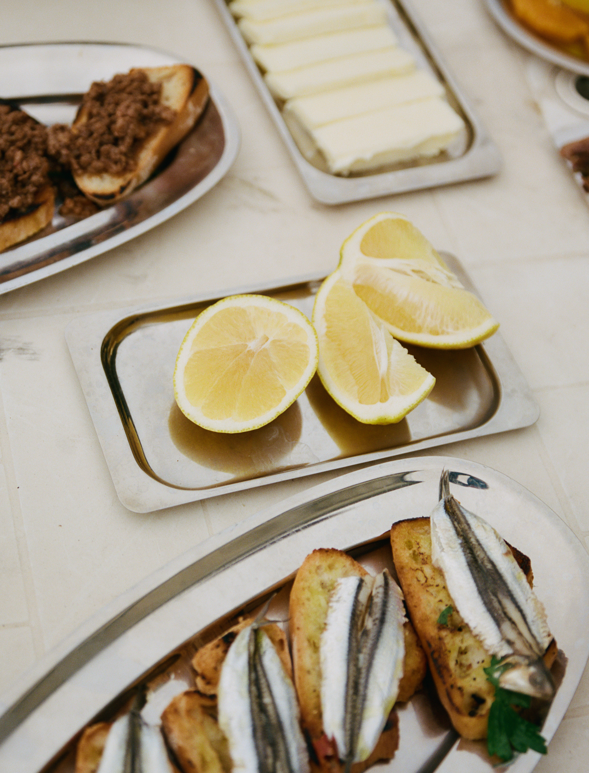 O16 Vassoio 3 kvadratisk stålfad med citronbåde blandt crostini og smør på et anretterbord — lifestyle | O16 Vassoio 3 square steel tray of lemon wedges among crostini and butter on a serving table — lifestyle shot