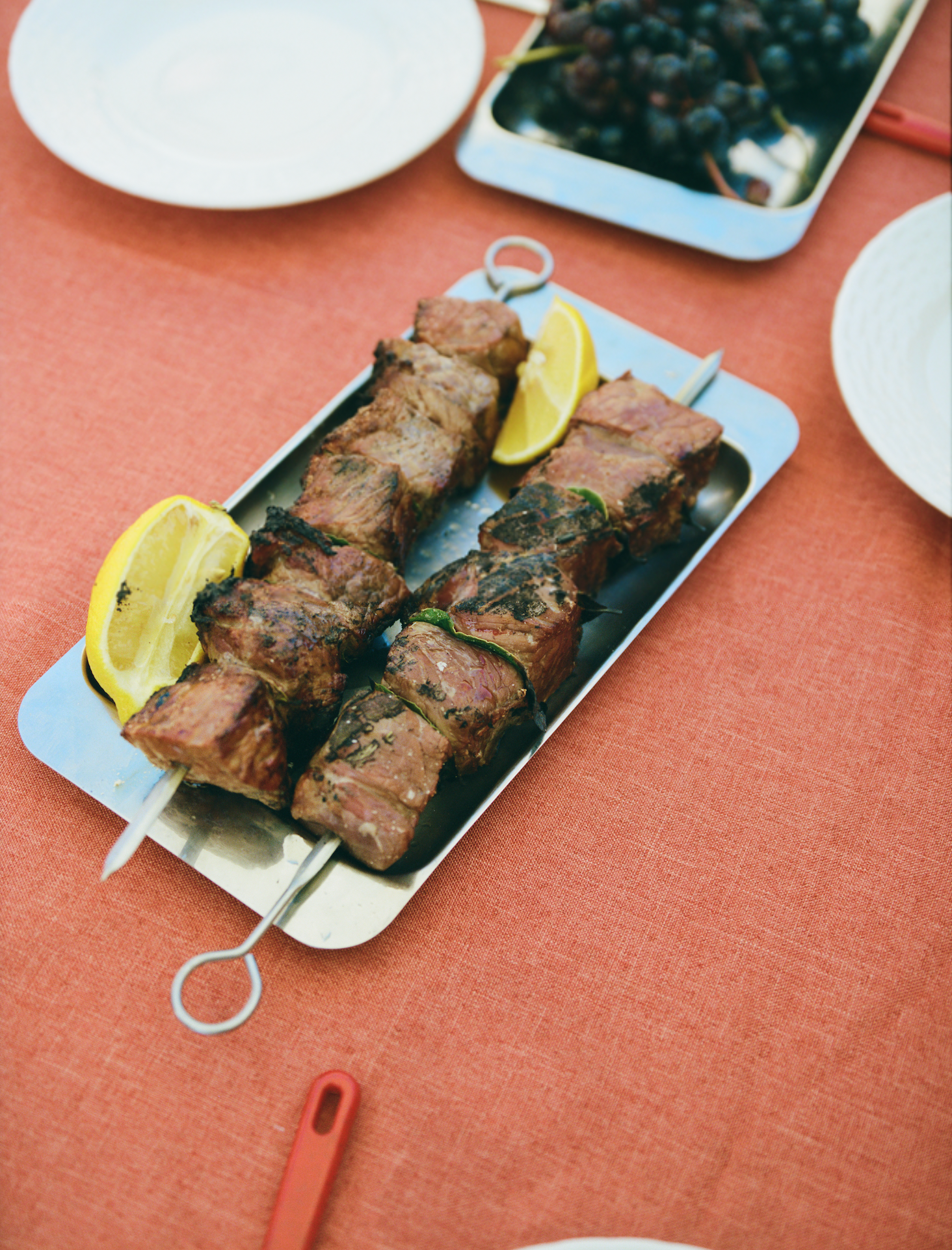 In-use: O16 Vassoio 4 rektangulært stålfad med grillede kødspyd og citron på koralrød dug — lifestyle | O16 Vassoio 4 rectangular steel tray with grilled meat skewers and lemon on a coral tablecloth — lifestyle shot
