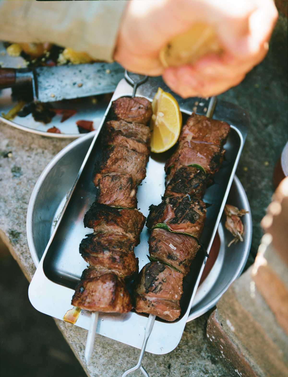 O16 Vassoio 4 rektangulært stålfad med grillede spyd og citron, hånd der presser citron over — lifestyle | O16 Vassoio 4 rectangular steel tray with grilled skewers and lemon, hand squeezing lemon over — lifestyle shot