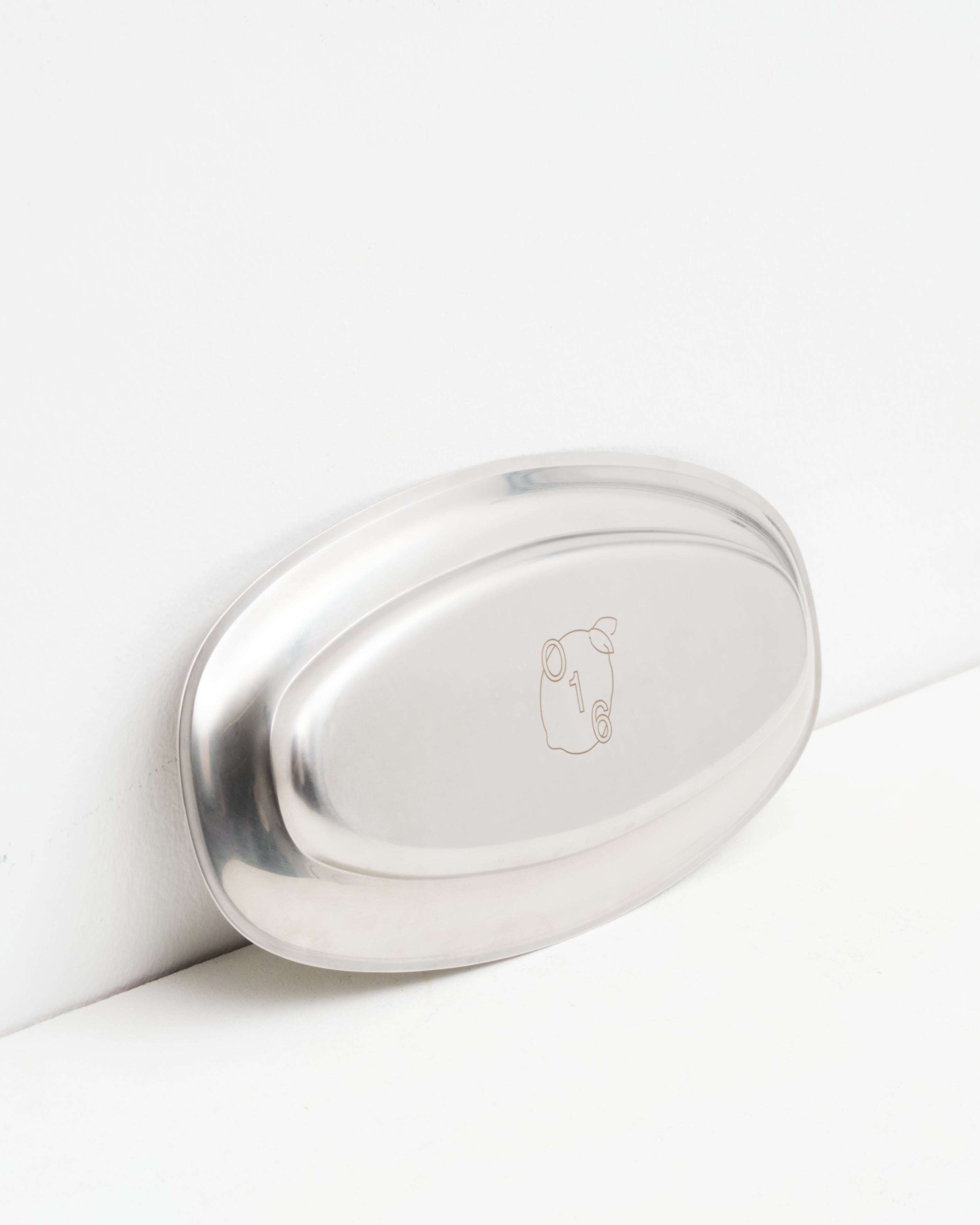 Studio: O16 Osteria 16 Vassoio 2 mellemstort ovalt serveringsfad i rustfrit stål lænet mod væg med indgraveret O16-logo, detaljebillede | O16 Osteria 16 Vassoio 2 medium oval stainless steel serving tray leaning against a wall with engraved O16 logo, detail shot