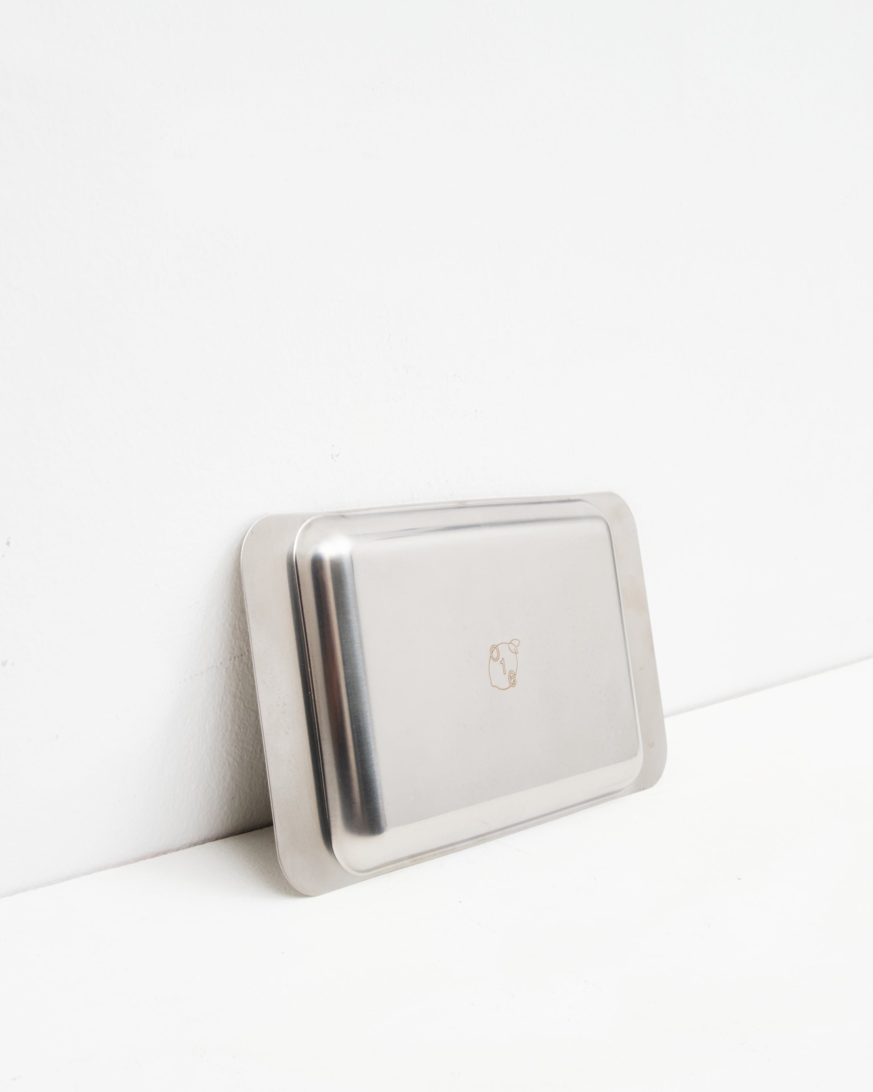 Studio: O16 Osteria 16 Vassoio 4 rektangulært serveringsfad i rustfrit stål lænet mod væg med indgraveret O16-logo, detaljebillede | O16 Osteria 16 Vassoio 4 rectangular stainless steel serving tray leaning against a wall with engraved O16 logo, detail shot