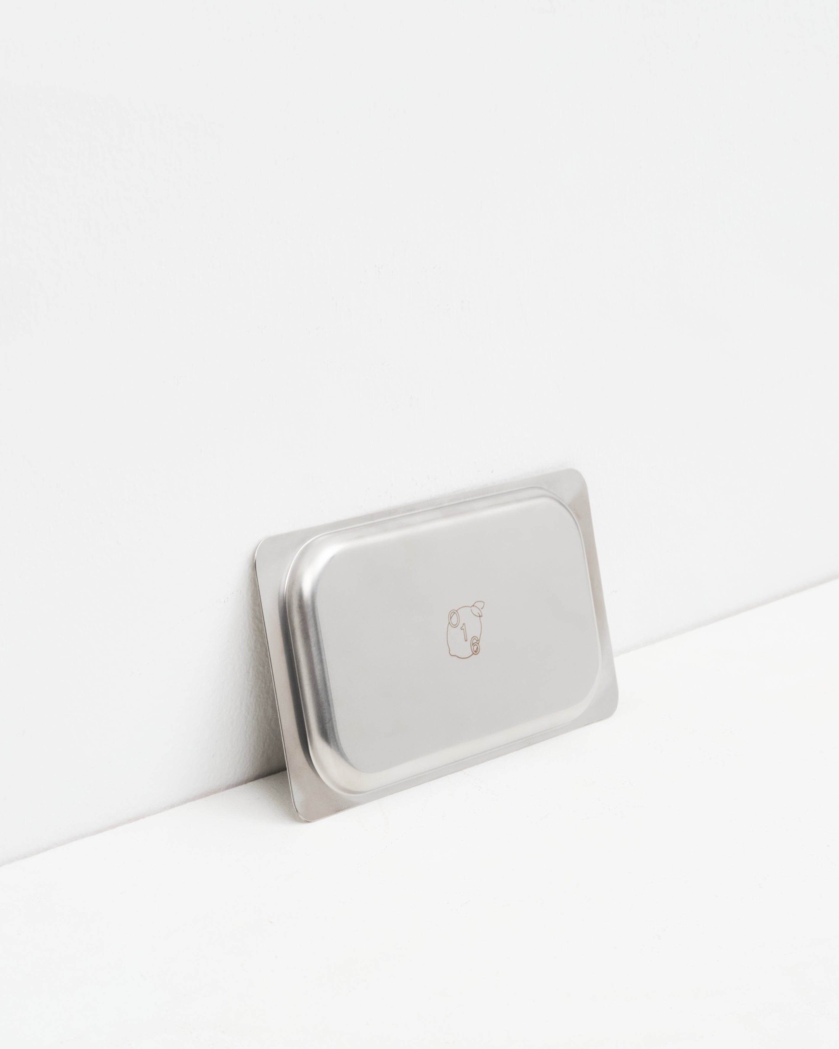 Studio: O16 Osteria 16 Vassoio 3 lille rektangulært serveringsfad i rustfrit stål lænet mod væg med indgraveret O16-logo, detaljebillede | O16 Osteria 16 Vassoio 3 small rectangular stainless steel serving tray leaning against a wall with engraved O16 logo, detail shot