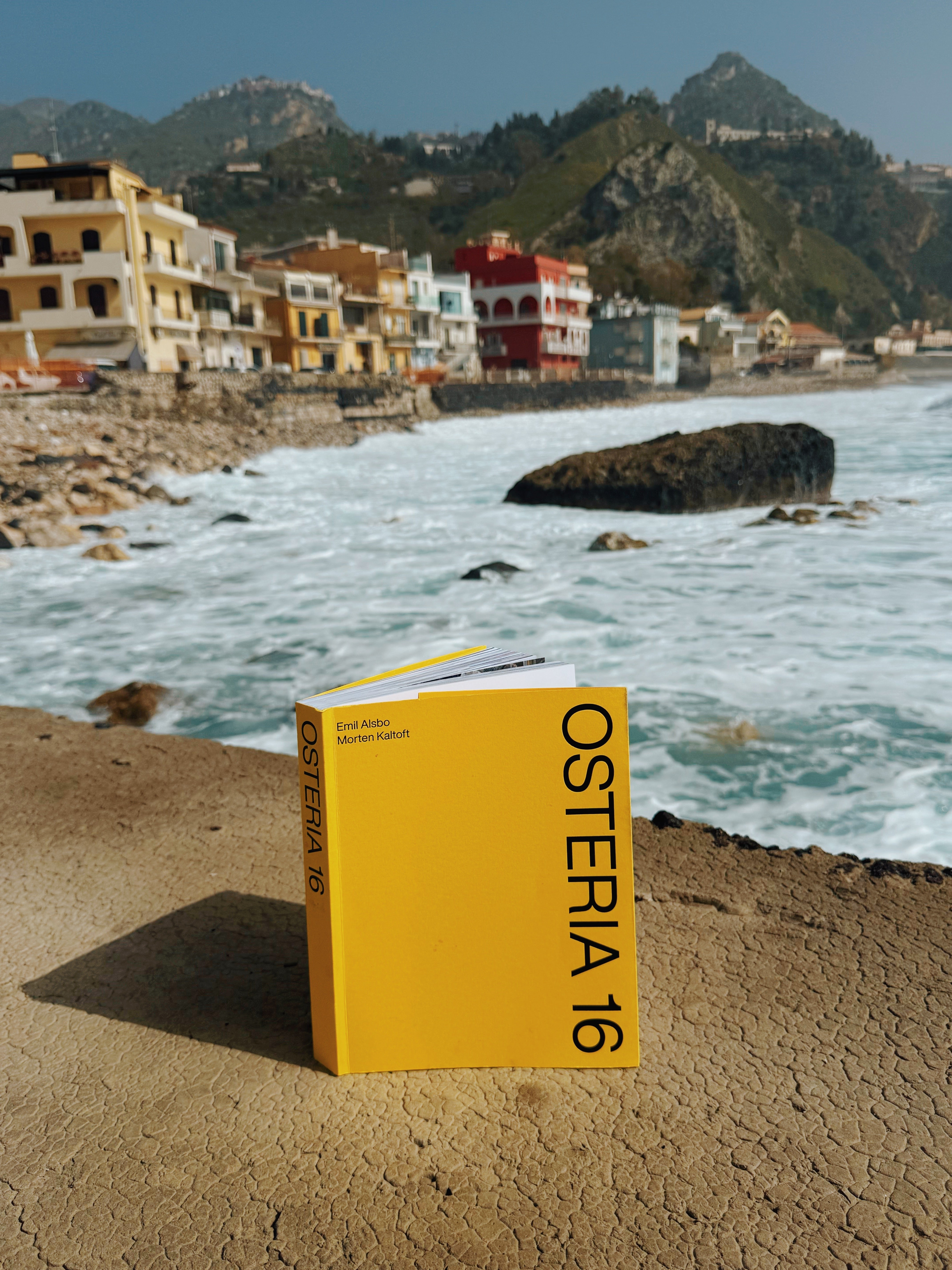In-use: Gul Osteria 16 kogebog af Emil Alsbo og Morten Kaltoft står på en havmur ved en siciliansk kystby. | Yellow Osteria 16 cookbook by Emil Alsbo and Morten Kaltoft standing on a seawall by a Sicilian coastal town.